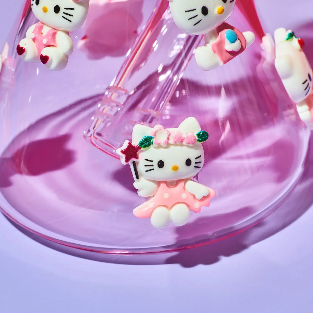 Hello Kitty character charms in a pink and white polka-dot dress, shown hanging from a clear glass rod inside a pink glass bowl on a purple background.