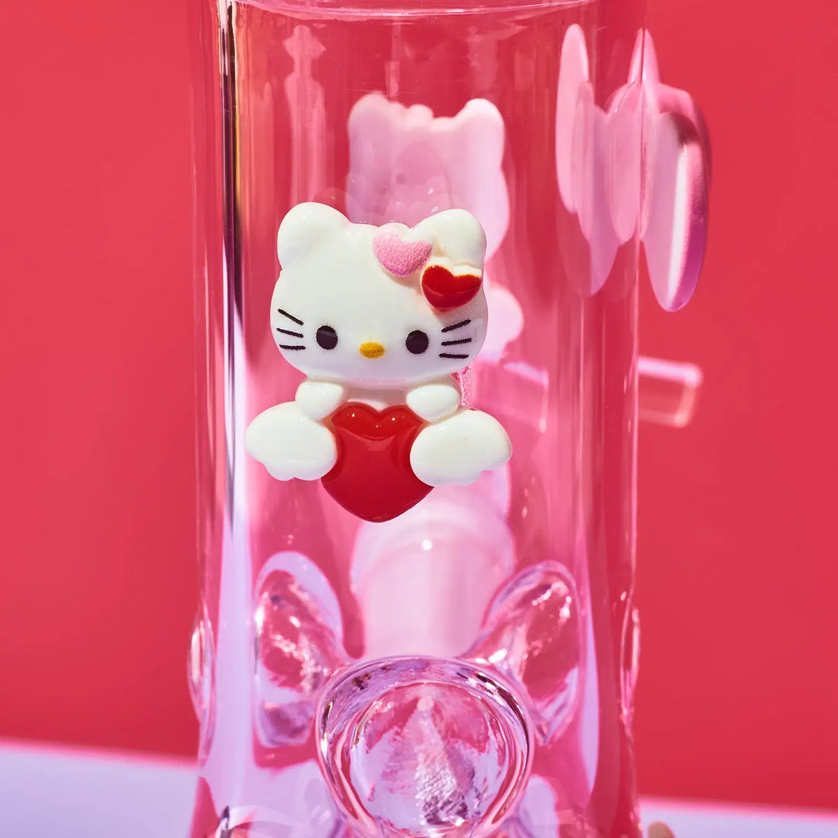 A pink transparent glass tumbler featuring a Hello Kitty character decoration holding a red heart, set against a solid pink background.