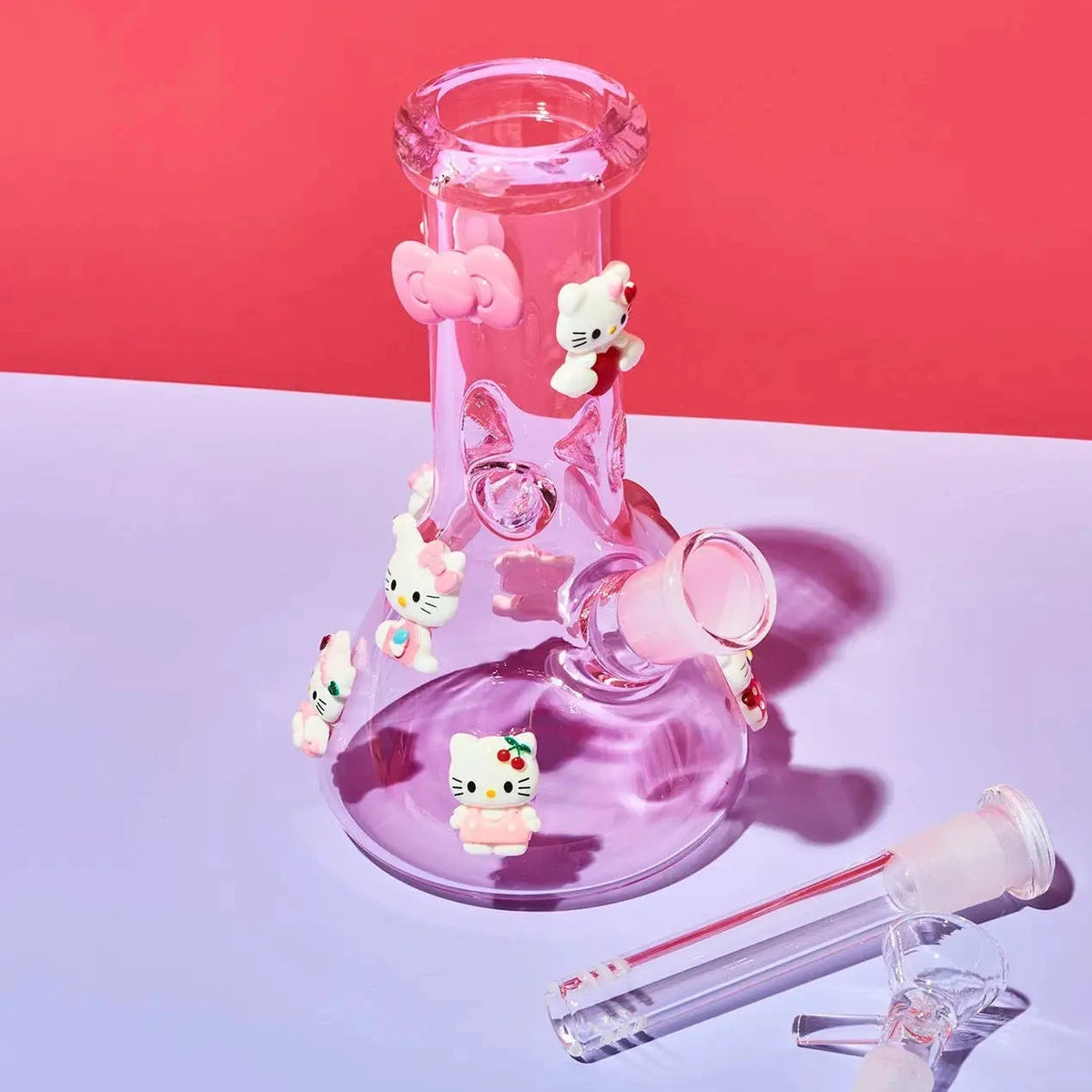 A pink glass water pipe with Hello Kitty character decorations, shown with its glass mouthpiece and bowl piece on a split pink and purple studio background.