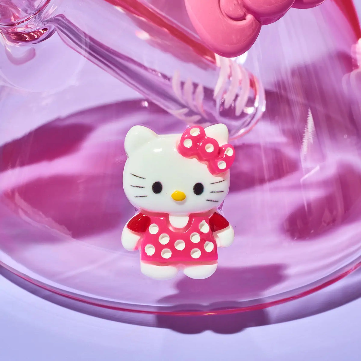 A small Hello Kitty character charm or figurine wearing a pink polka-dot dress and matching bow, posed inside a translucent pink glass container.