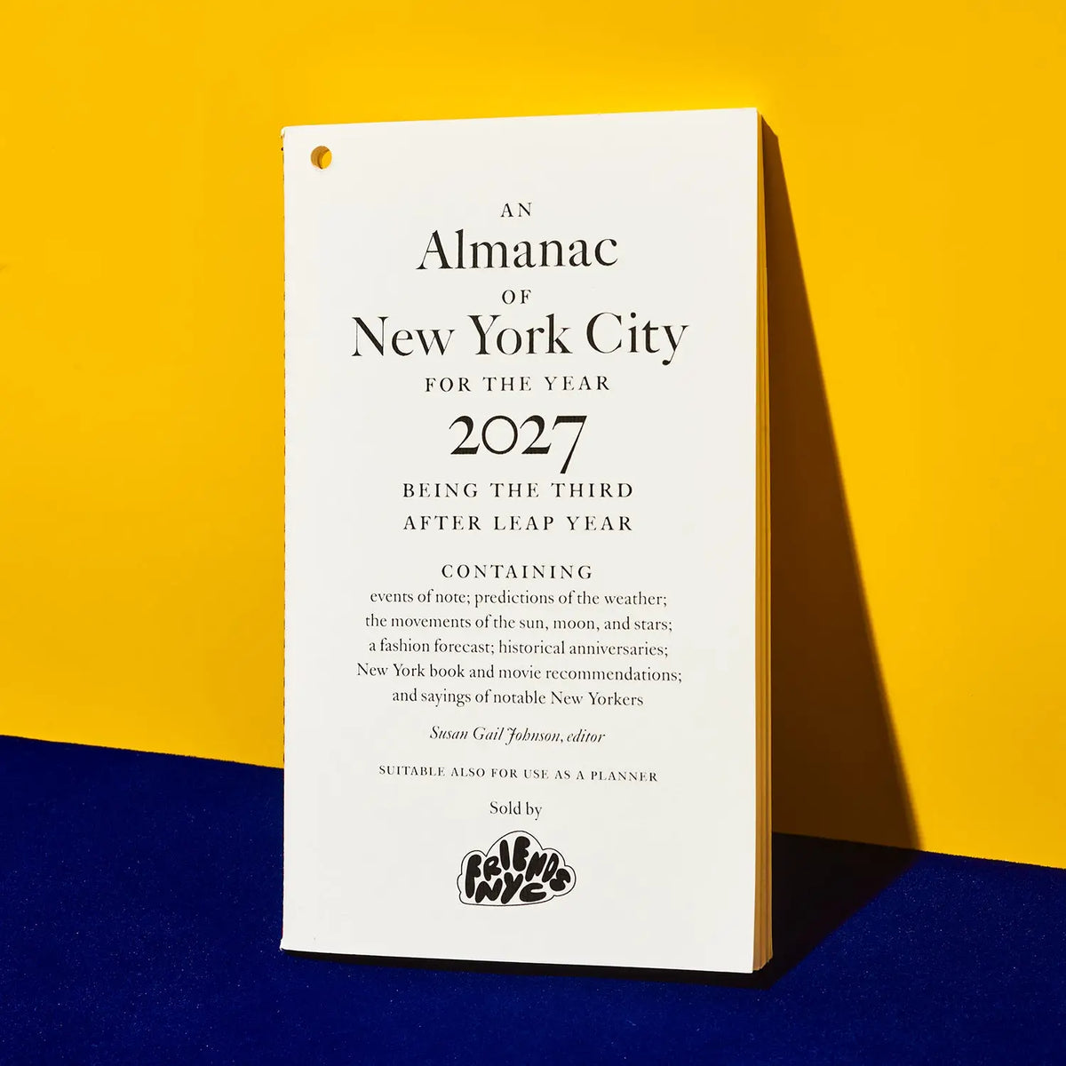 A tall, narrow white planner or calendar cover standing upright against a two-tone yellow and dark blue background. The front cover features black serif text reading 'An Almanac of New York City for the Year 2027' and smaller descriptive text below, with a small black logo near the bottom.