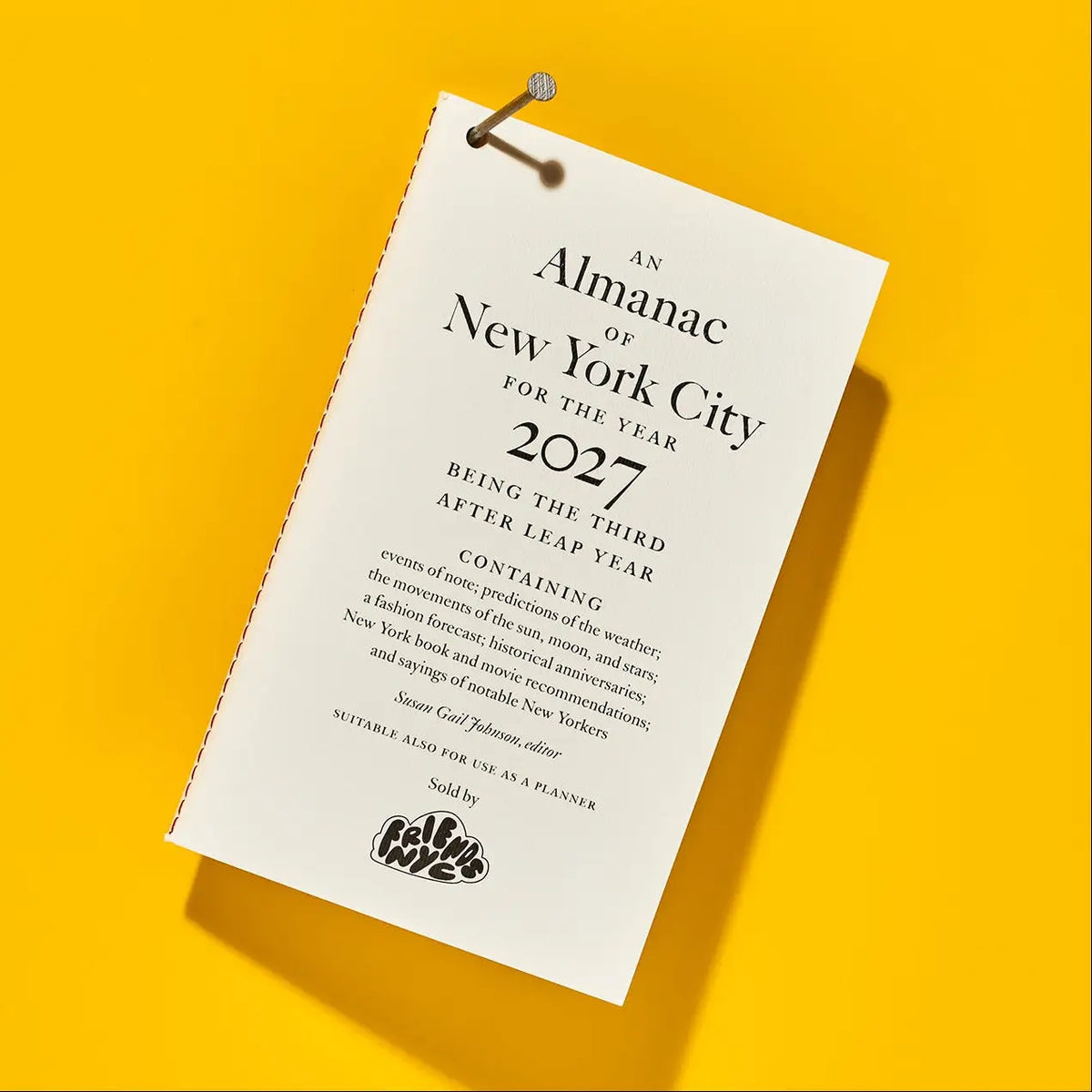 A single white paper planner or calendar page with printed text reading 'An Almanac of New York City for the Year 2027' and a small logo at the bottom, photographed on a bright yellow background with a pin holding it.