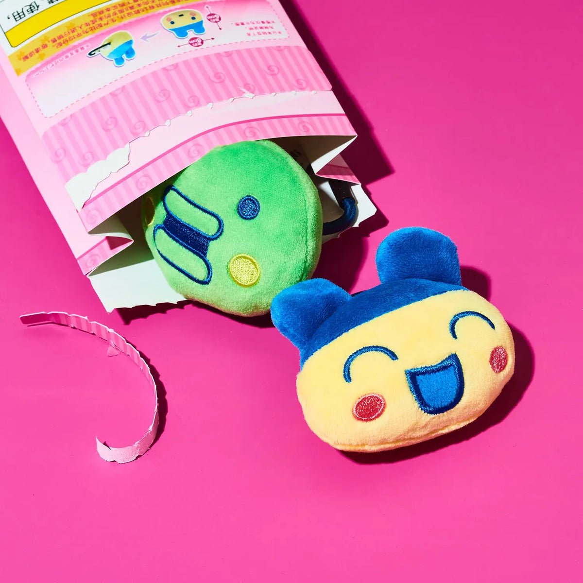 A plush emoji face toy in a yellow and blue design, shown next to its pink packaging on a bright pink background.