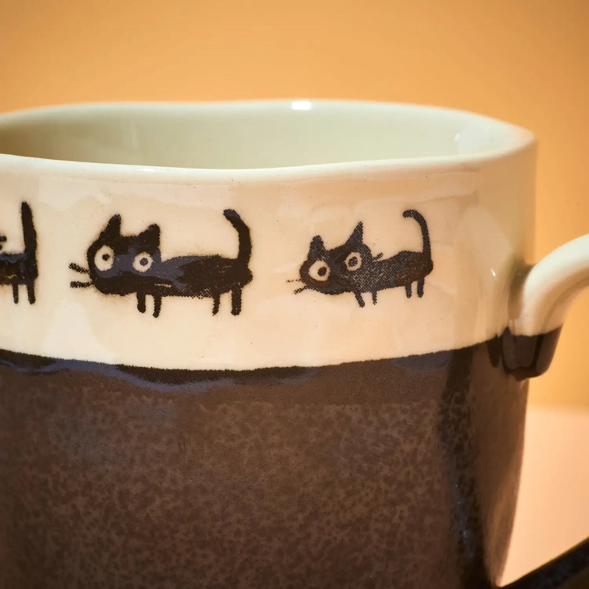 Close-up of a ceramic mug with a white upper band decorated with black hand-drawn cat silhouettes, and a dark brown speckled lower body. A white handle is visible on the right against a warm beige background.