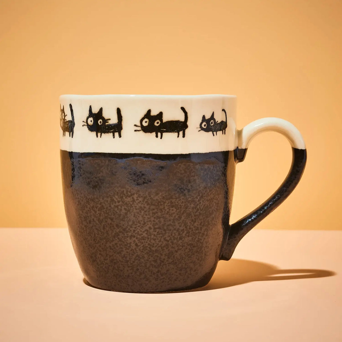 A ceramic mug with a speckled dark brown body, a white band decorated with black cat silhouettes, and a white handle, set against a warm beige background.