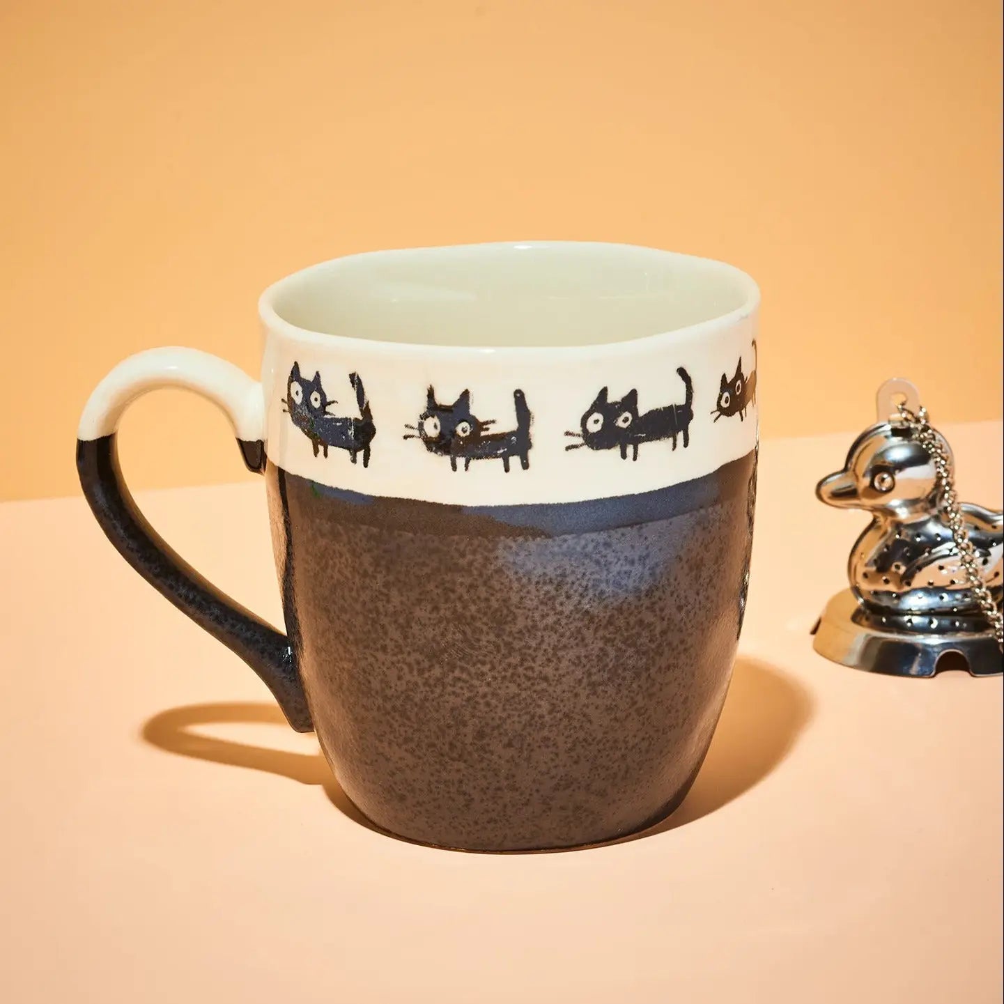 A ceramic mug with a speckled dark brown lower half, a white upper band decorated with black cat illustrations, and a black handle, shown on a peach-colored background next to a small silver duck figurine.