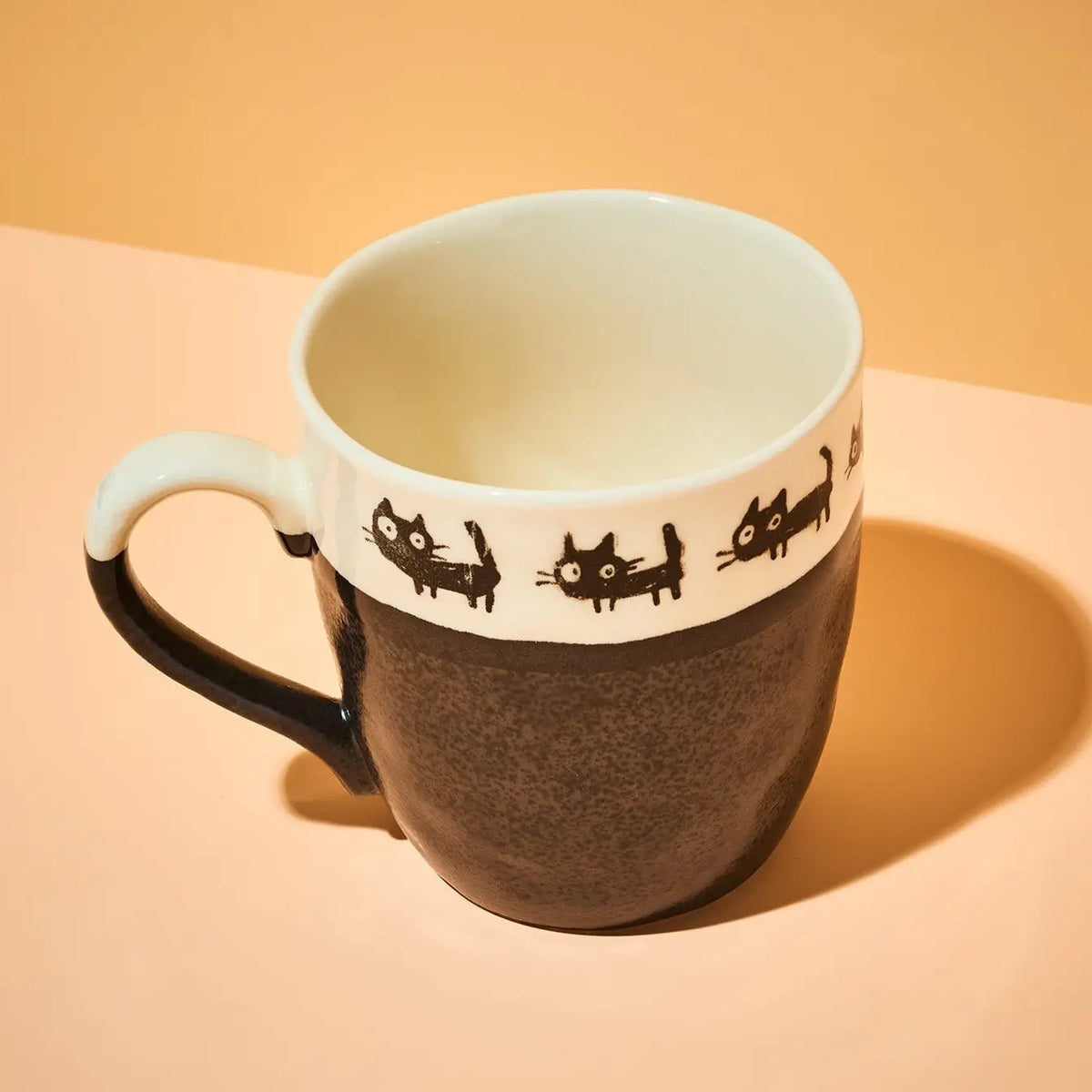 A ceramic mug with a dark brown speckled body, a white band decorated with black cat illustrations, and a black handle, set against a warm peach and beige background.