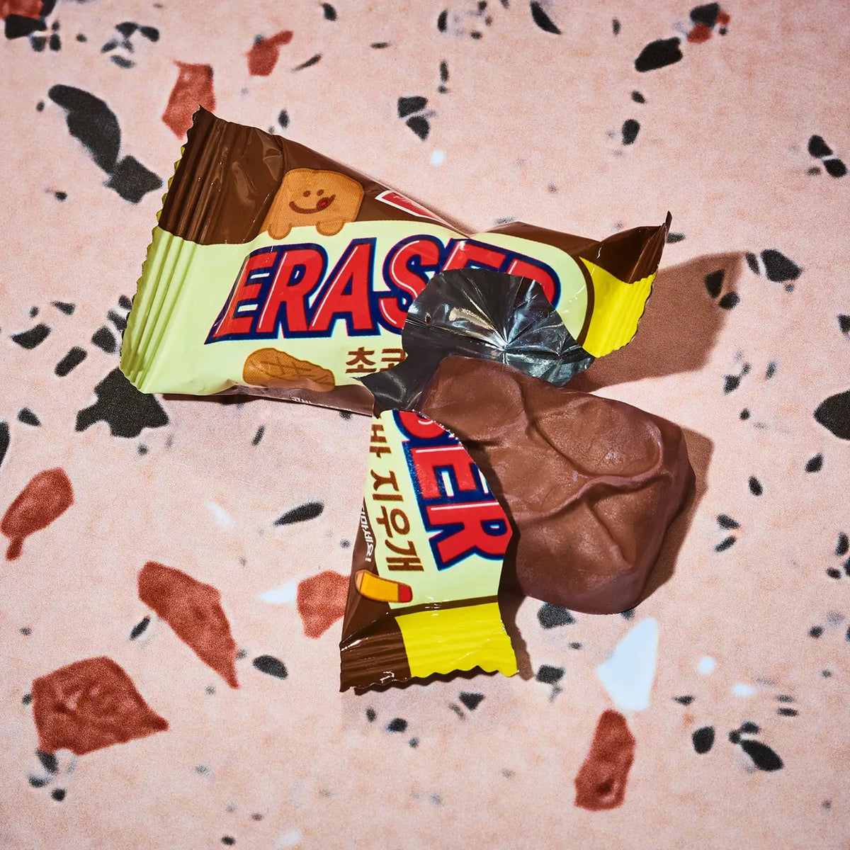 A close-up of a partially unwrapped Snickers ERASED chocolate bar, showing the brown chocolate coating and the cream-colored wrapper with red and blue lettering, placed on a pink terrazzo surface.