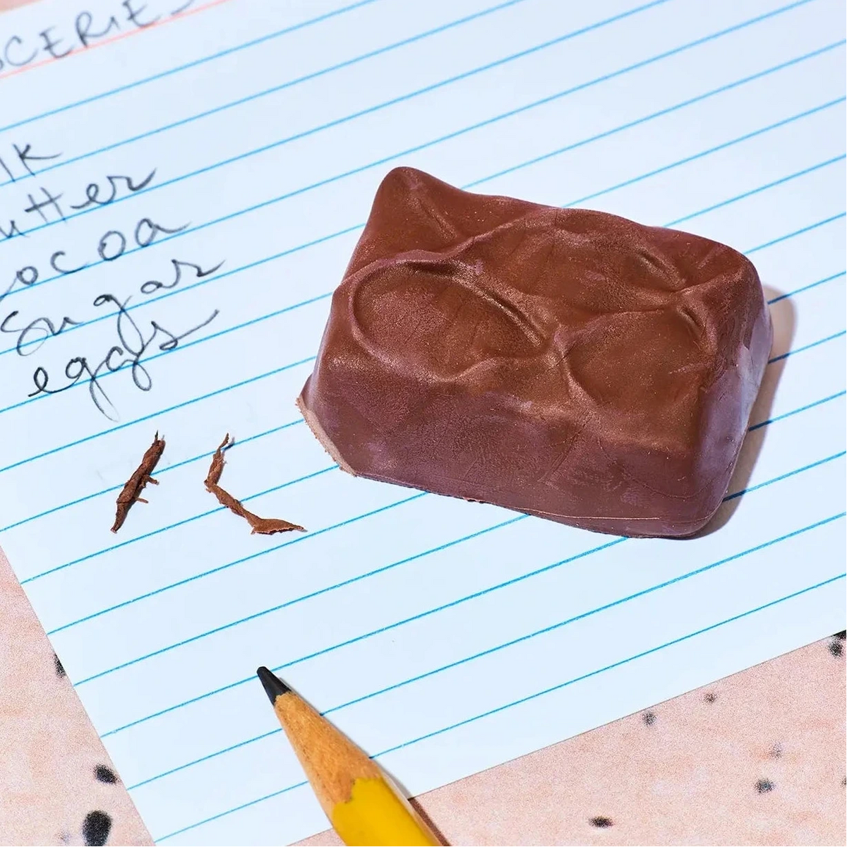 A realistic replica of a milk chocolate bar with a textured top, placed on a piece of lined notebook paper next to a yellow pencil and handwritten notes spelling out ingredients.