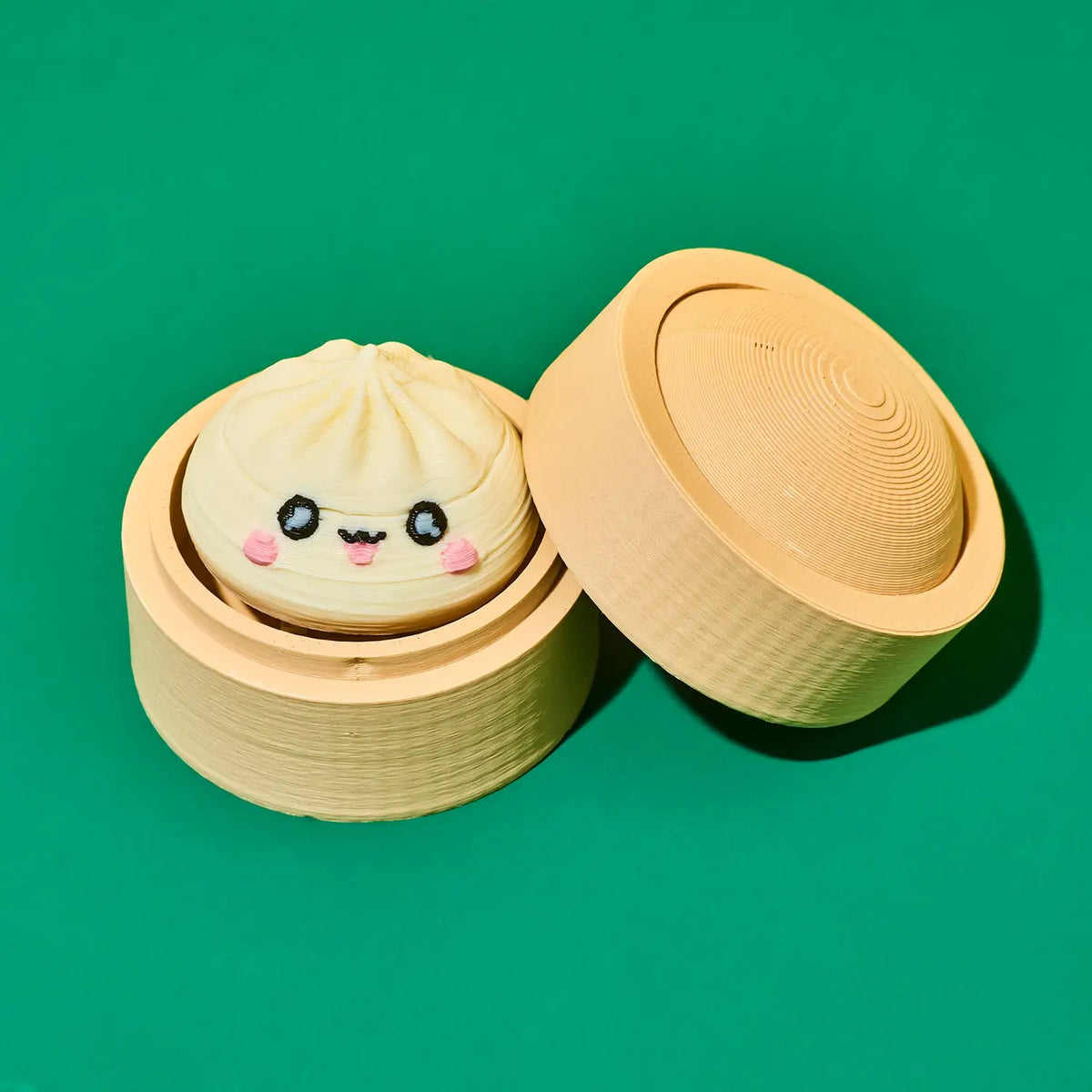 A cute, kawaii-style mini steamer basket toy or container featuring a small embroidered dumpling character with a smiling face and pink cheeks, shown alongside its lid on a solid green background.