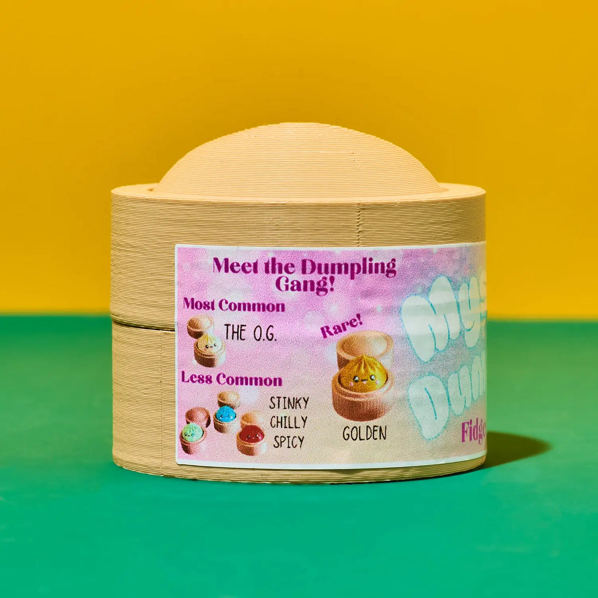 A small round wooden container with a domed lid, shown on a bright yellow and green background. The front has a colorful printed label reading 'Meet the Dumpling Gang!' with illustrations of dumplings and text including 'Most Common', 'Rare!', 'Stinky Chilly Spicy', and 'Golden'.