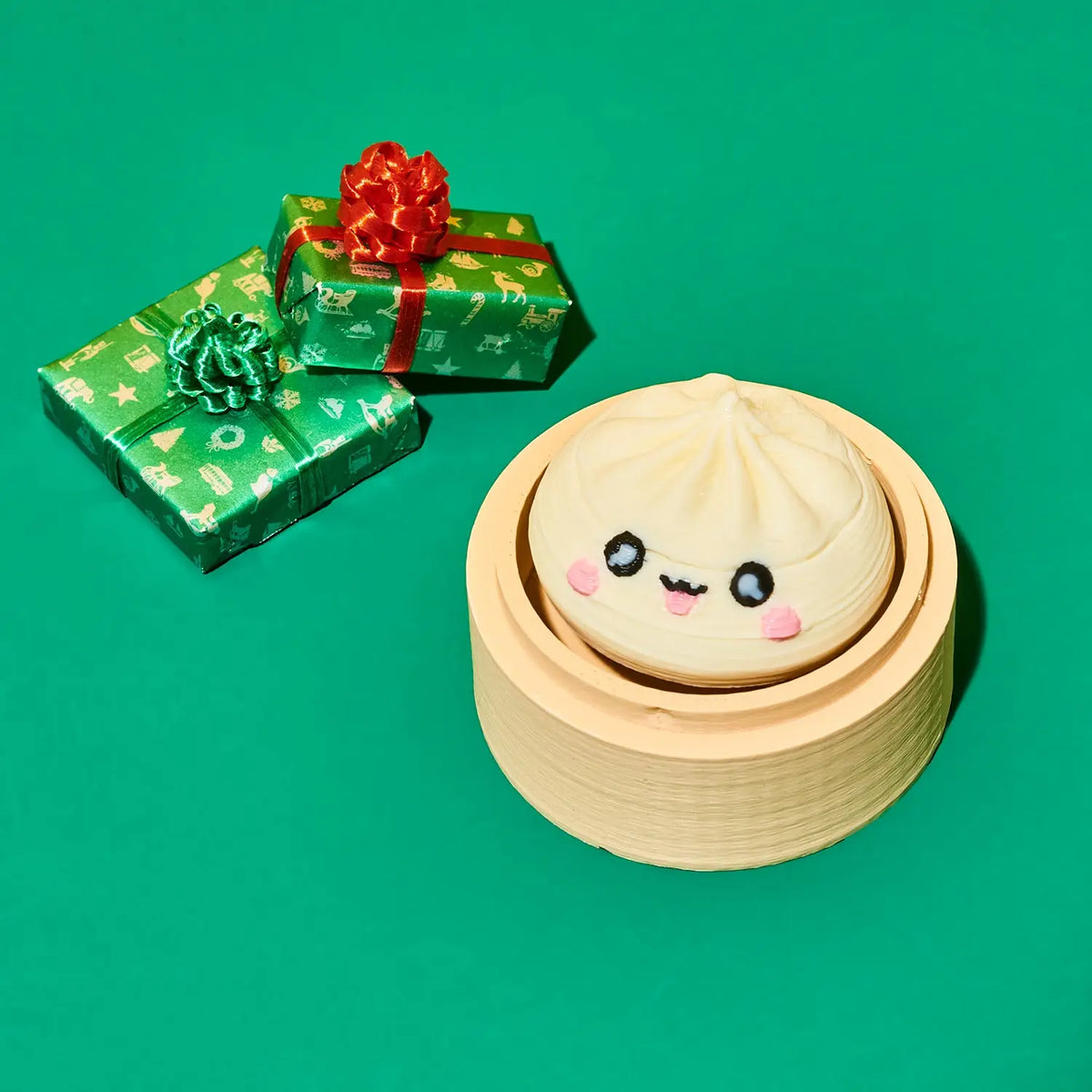 A cute, kawaii-style dumpling character with a smiling face sits inside a round bamboo steamer basket on a solid green background. Two wrapped gift boxes with green patterned paper and red bows are placed in the background.