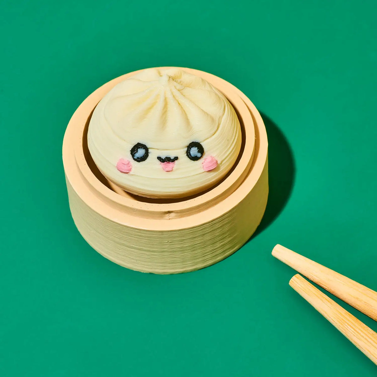 A cute, round steamed bun decoration or toy with a smiling face, sitting inside a wooden steamer basket on a solid green background with wooden chopsticks in the corner.
