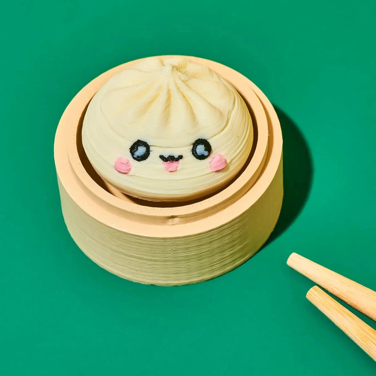 A cute, round steamed bun decoration or toy with a smiling face, sitting inside a wooden steamer basket on a solid green background with wooden chopsticks in the corner.