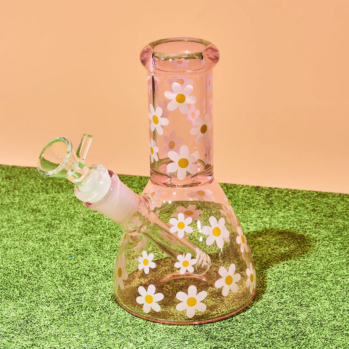 A pink glass water pipe with a white and yellow daisy pattern, resting on green artificial grass against a peach background.