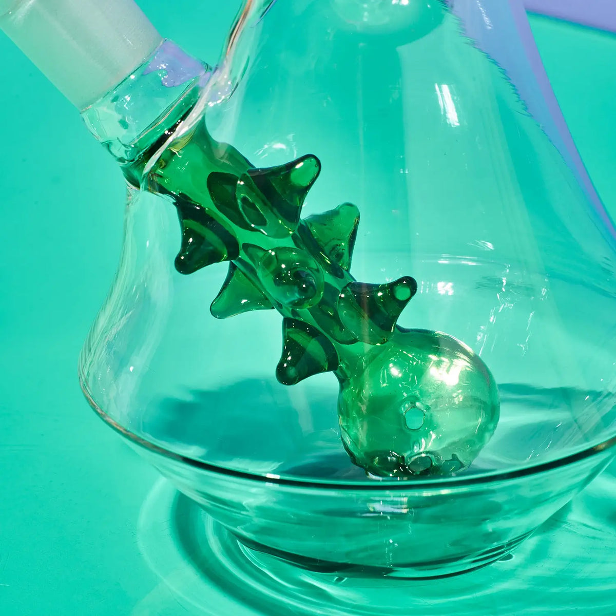 A close-up of a transparent glass vessel with a sculptural green glass spout and a matching green glass bulb, set against a bright turquoise background.