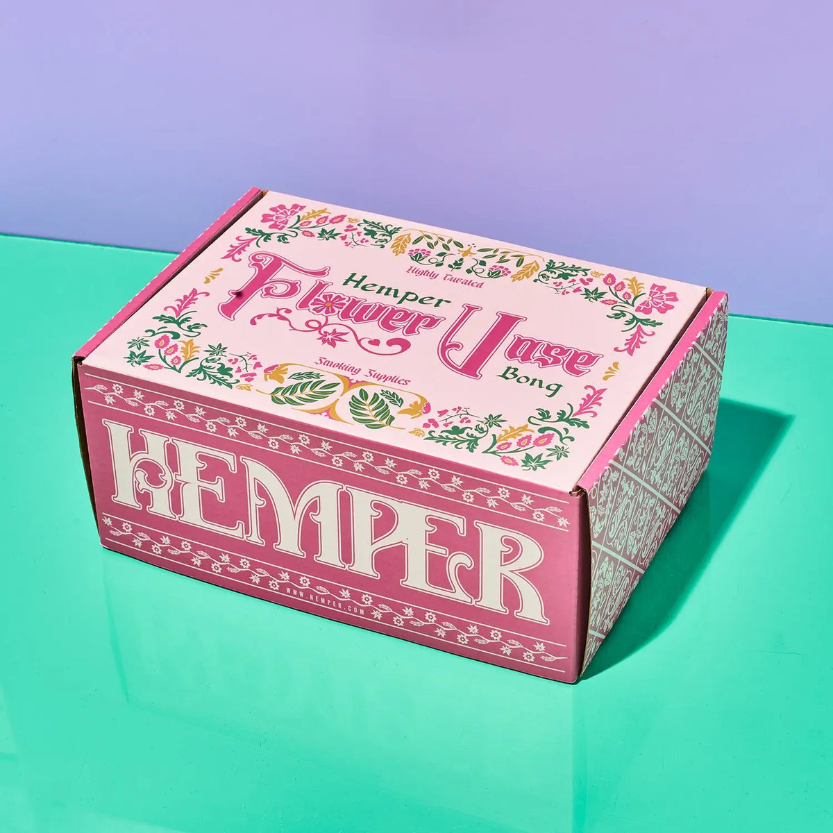 A pink cardboard box for Hemper Flower Vase Bong, photographed on a reflective mint-green surface against a lavender background.