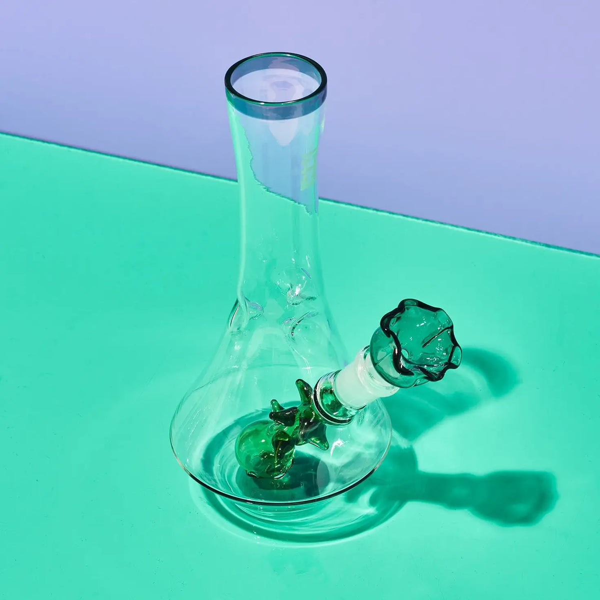 A clear glass water pipe with a green glass bowl and neck, set on a mint green surface against a lavender background.