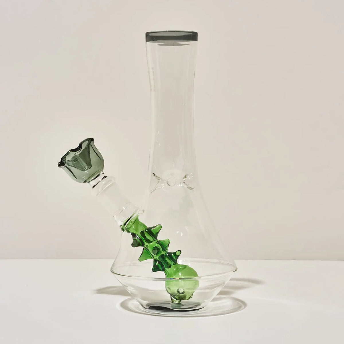 A clear glass water pipe or beaker-style glass container with a short neck, thick rounded base, and green glass bowl and bubble details, photographed on a white surface against a light neutral background.