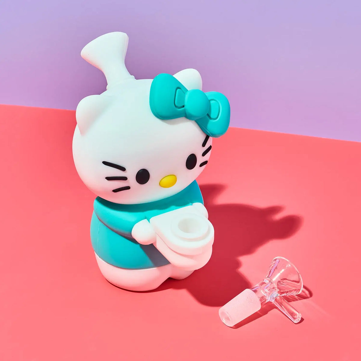 A Hello Kitty-shaped vape device in white and teal, shown with a clear glass mouthpiece accessory on a pink and purple studio background.