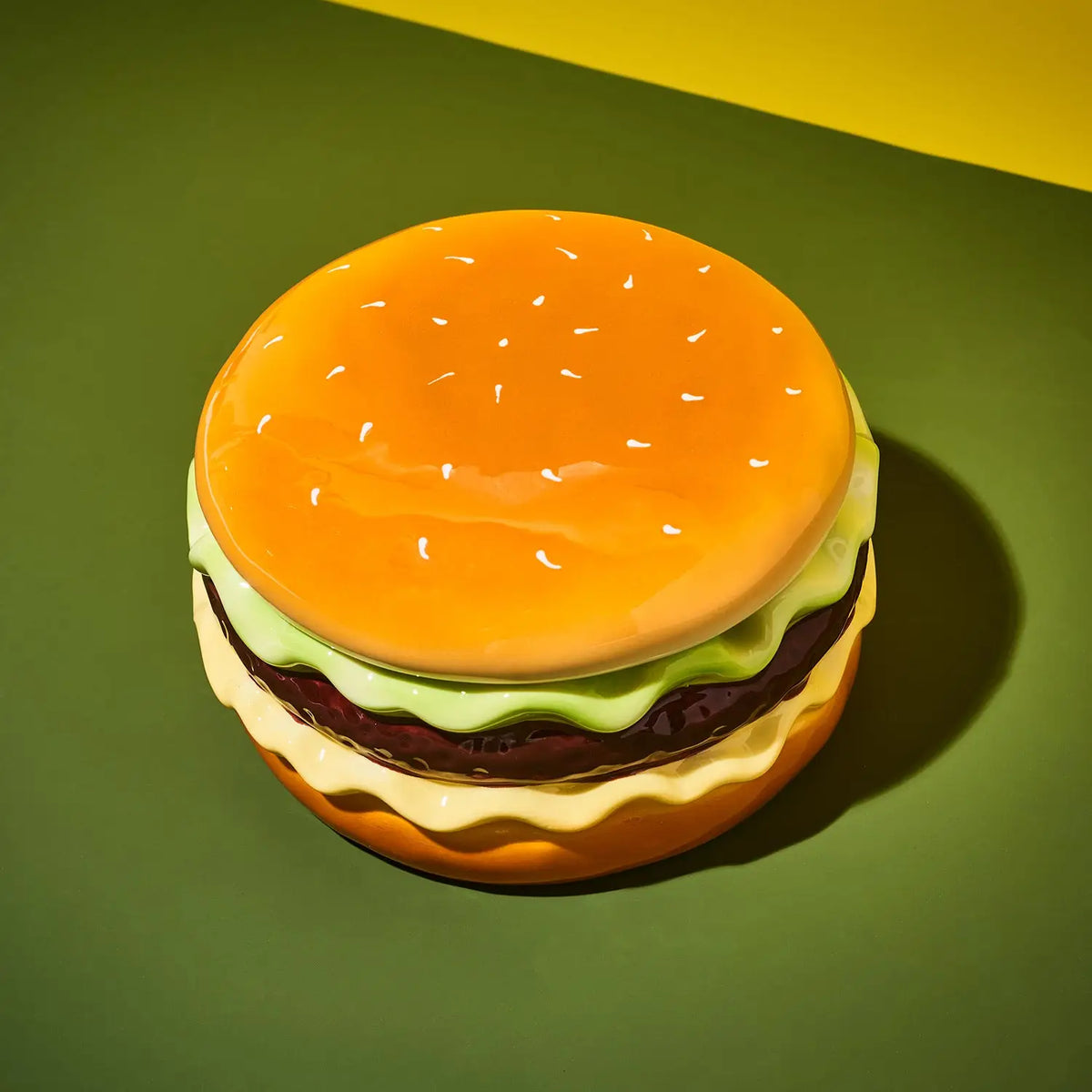 A glossy, stylized hamburger figurine or container on a two-tone green and yellow background.