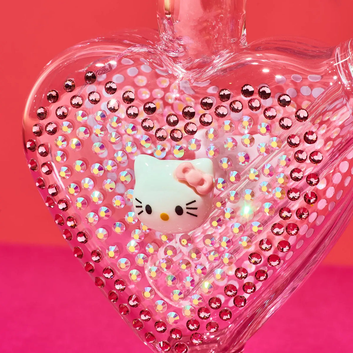 A heart-shaped clear glass bottle or ornament covered in pink rhinestones, featuring a white Hello Kitty face with a pink bow on the front, set against a bright pink background.