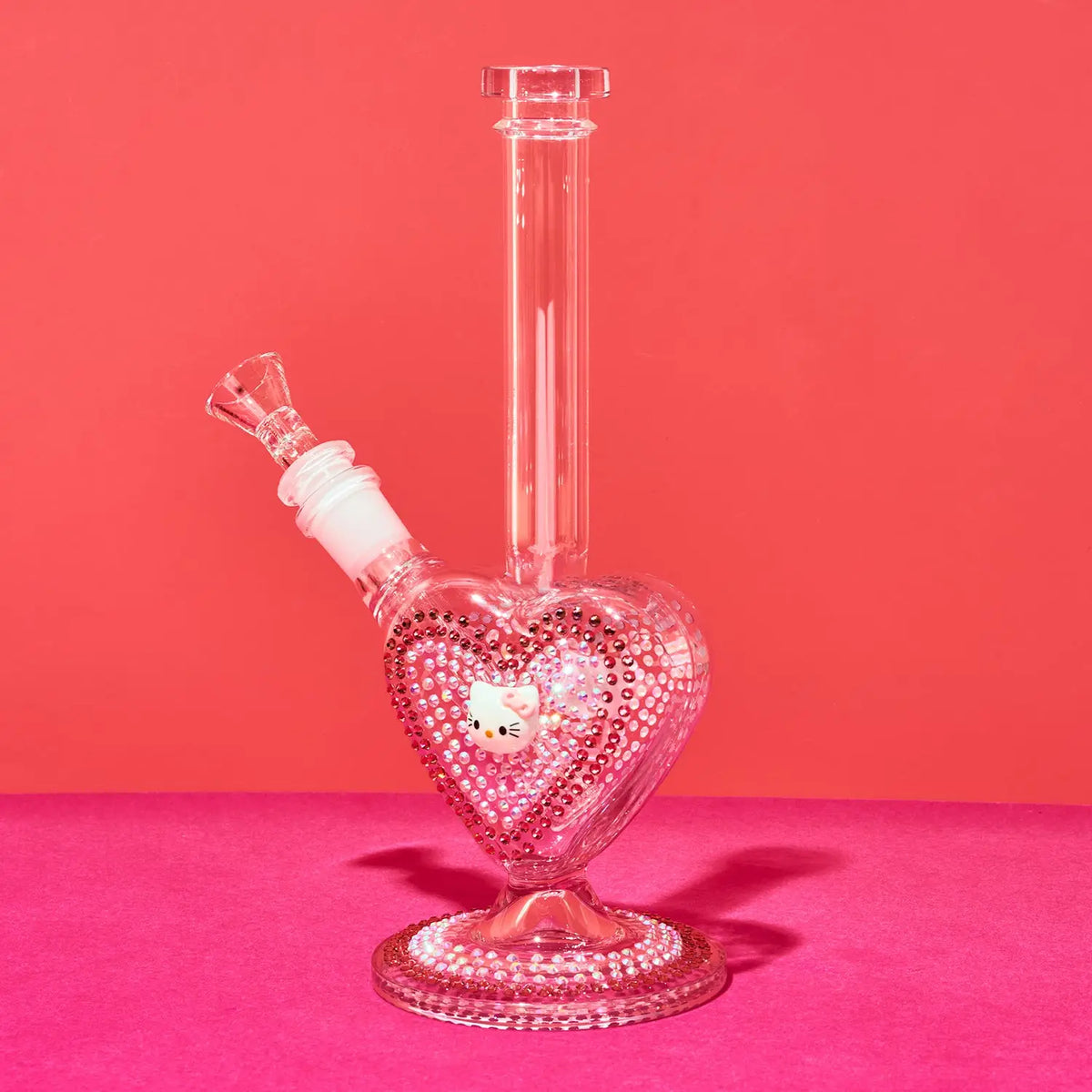 A clear glass water pipe with a heart-shaped base covered in pink rhinestones, featuring a Hello Kitty face charm in the center and a matching pink rhinestone-studded neck, set against a solid pink background.