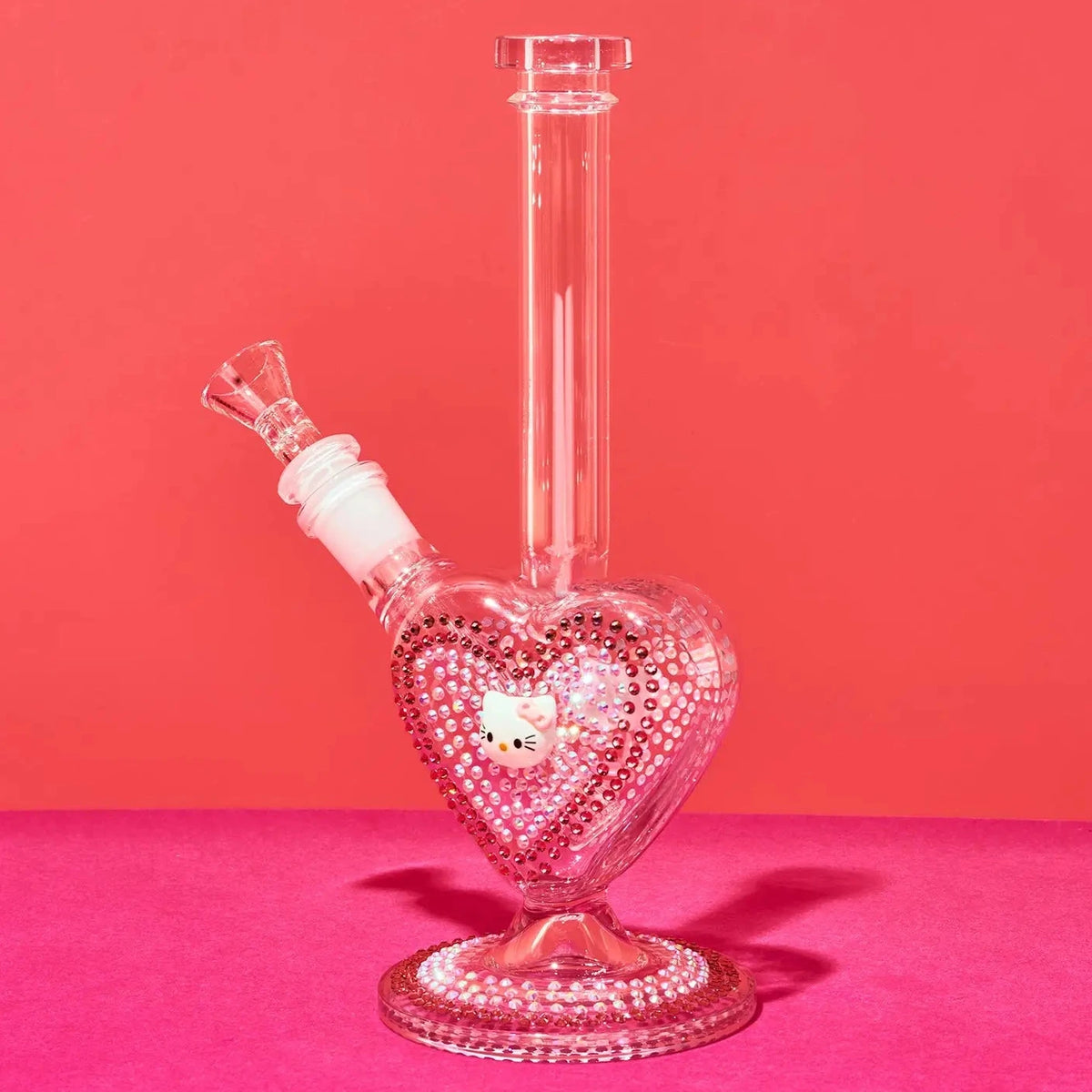 A clear glass water pipe with a heart-shaped base covered in pink rhinestones, featuring a Hello Kitty face charm in the center and a matching pink rhinestone-studded neck, set against a solid pink background.