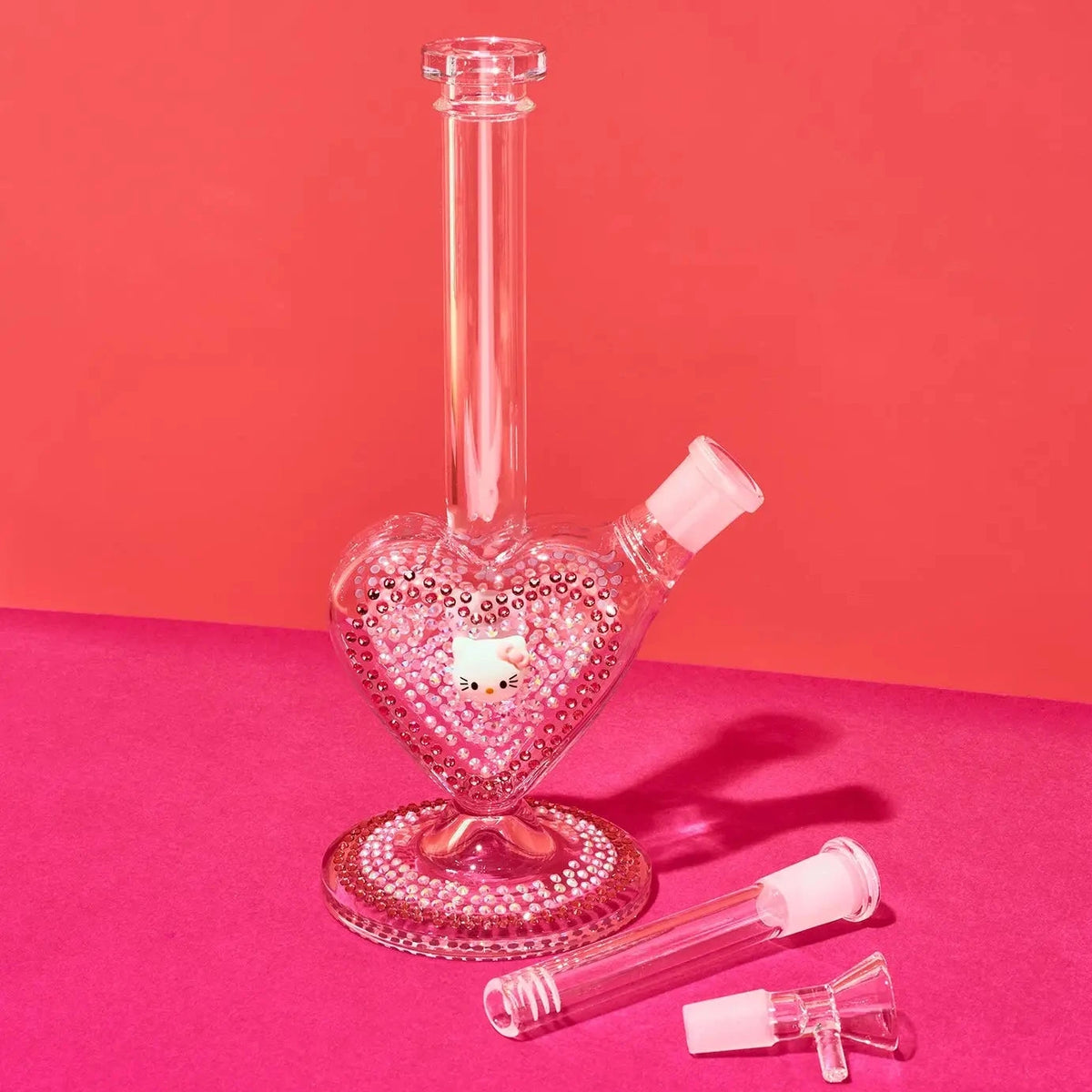 A pink heart-shaped glass water pipe with a long neck and a small side bowl, shown on a bright pink and coral background with two detachable glass mouthpieces in front.