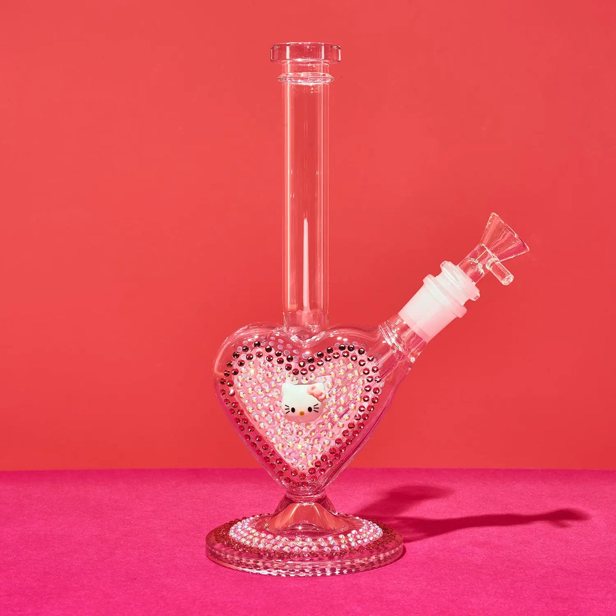 A clear glass water pipe with a heart-shaped base decorated with pink rhinestones and a small Hello Kitty figure inside, set against a solid pink background.