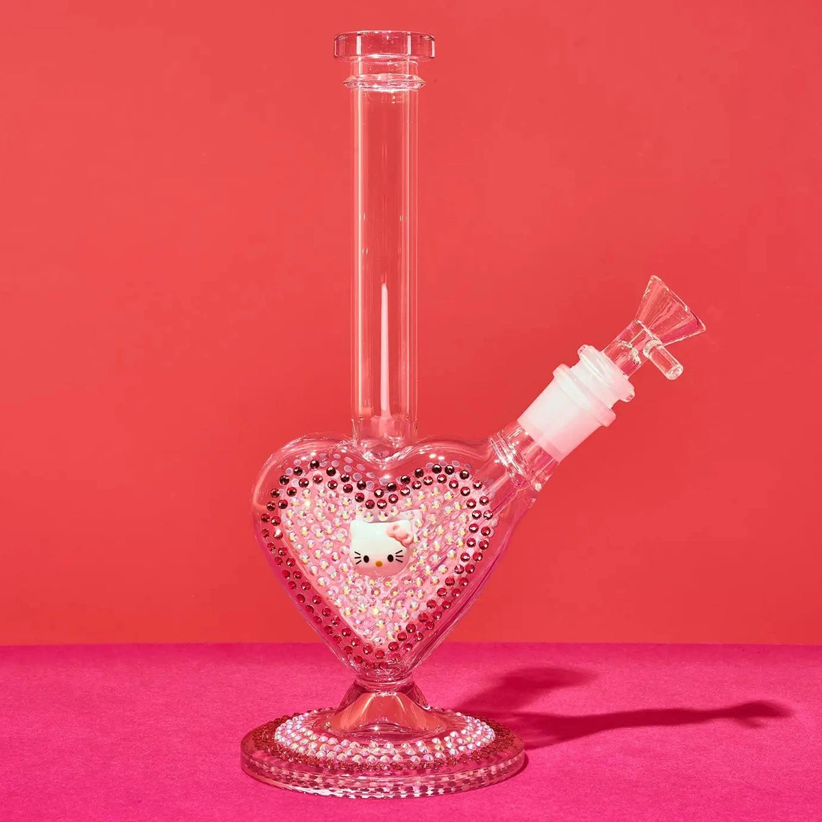 A clear glass water pipe with a heart-shaped base decorated with pink rhinestones and a small Hello Kitty figure inside, set against a solid pink background.