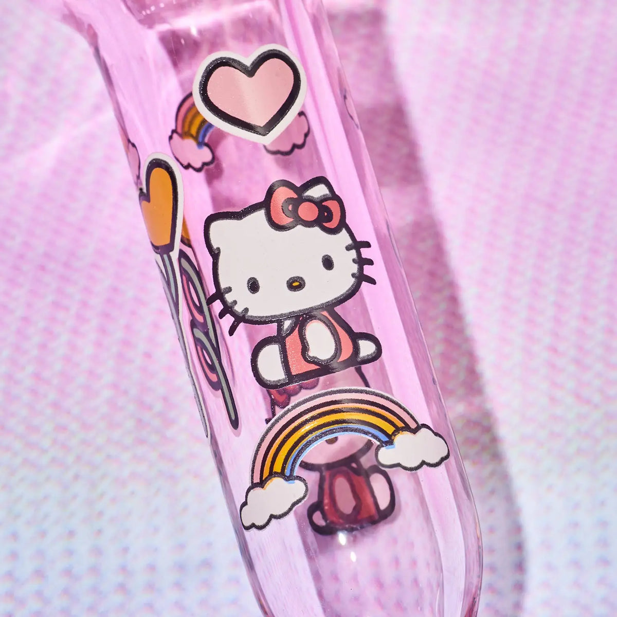 A close-up of a pink glass hand pipe decorated with Hello Kitty illustrations, including a heart, rainbow, clouds, and a pink bow, set against a textured pink background.