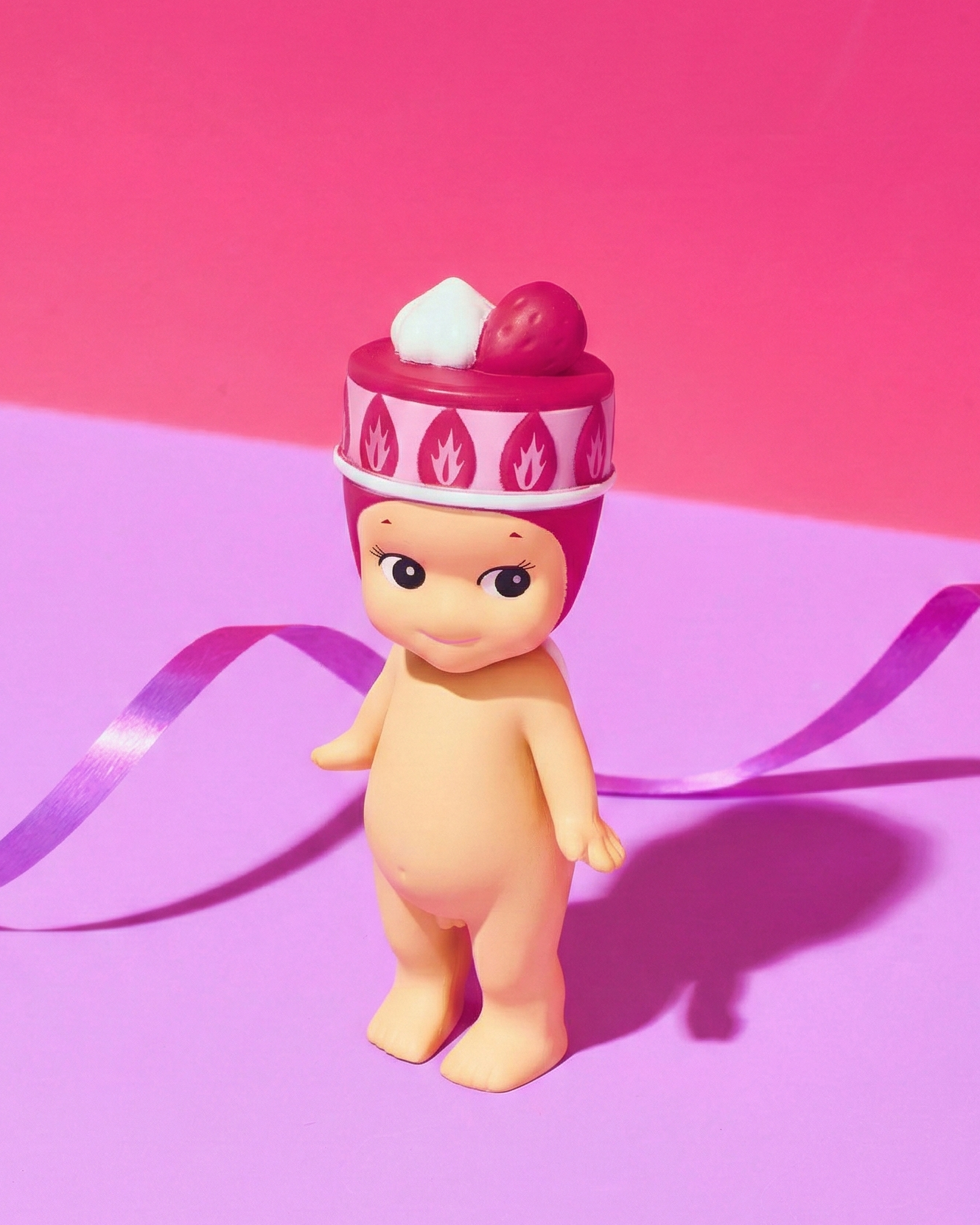 Toy figure with a red hat on a pink cake hat on a two tone background