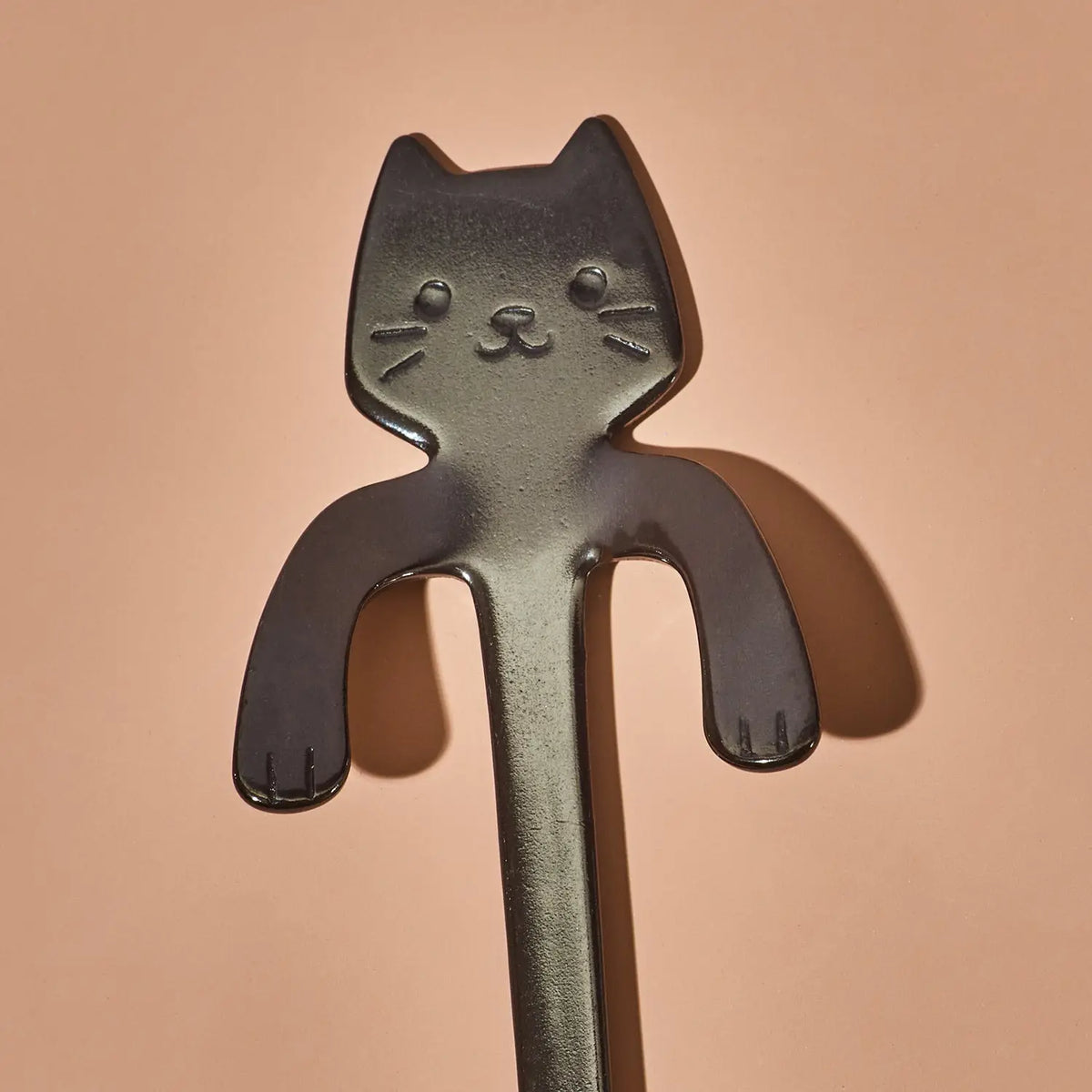 A dark gray metal cat-shaped utensil, likely a spoon or scoop, shown against a solid peach-colored background.