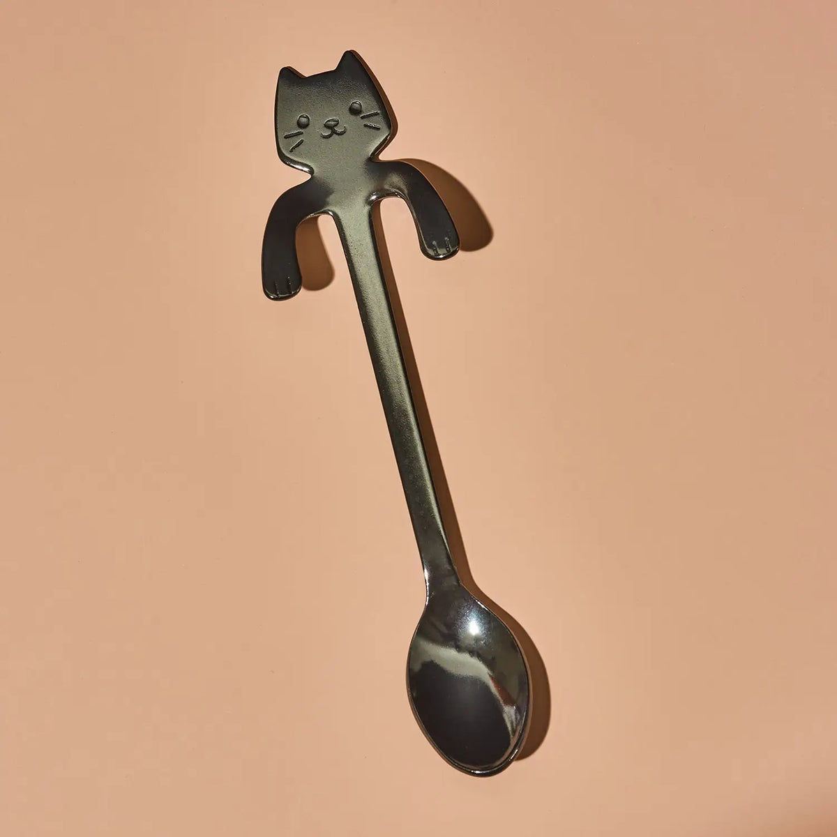 A novelty metal spoon with a cat-shaped handle, shown on a solid peach-colored background.