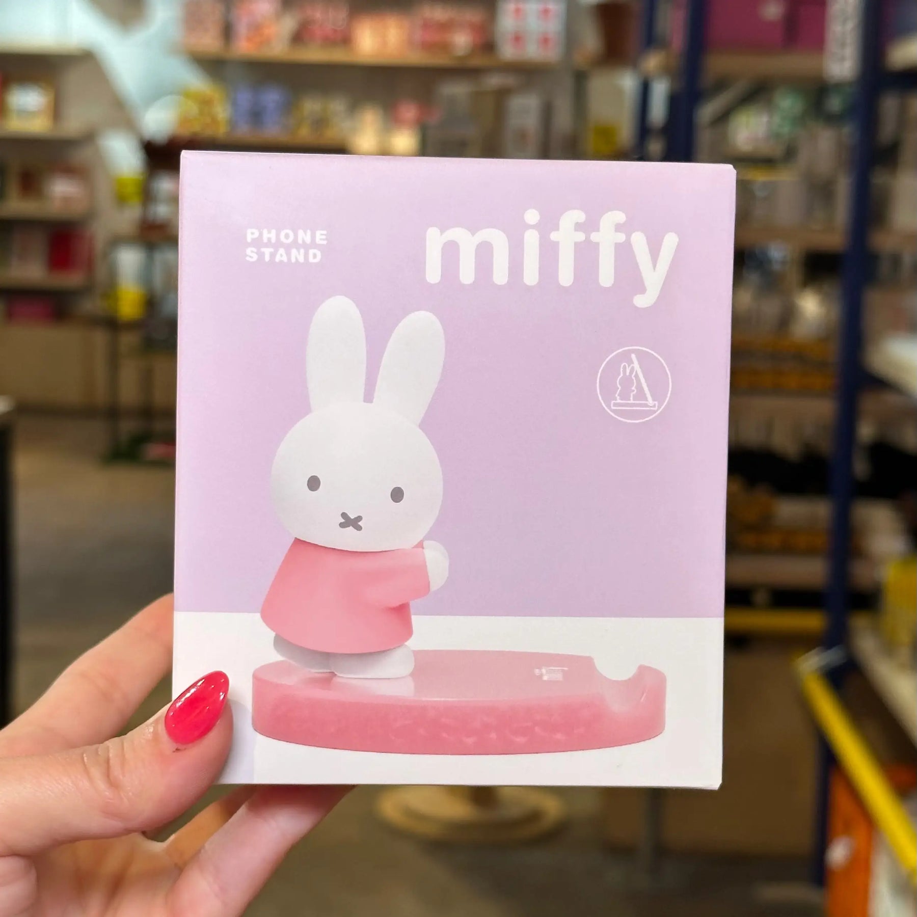 A hand with pink nail polish holds a light pink and white box for a Miffy phone stand in a retail store. The packaging features the character Miffy standing on a pink base, with the text 'PHONE STAND', 'miffy', and a circular logo.