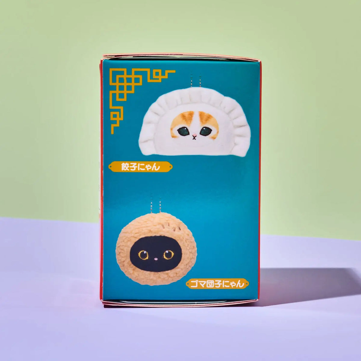 A boxed product standing upright on a pale lavender surface against a light green background. The package is teal with illustrated cat-faced dumplings and Japanese text.
