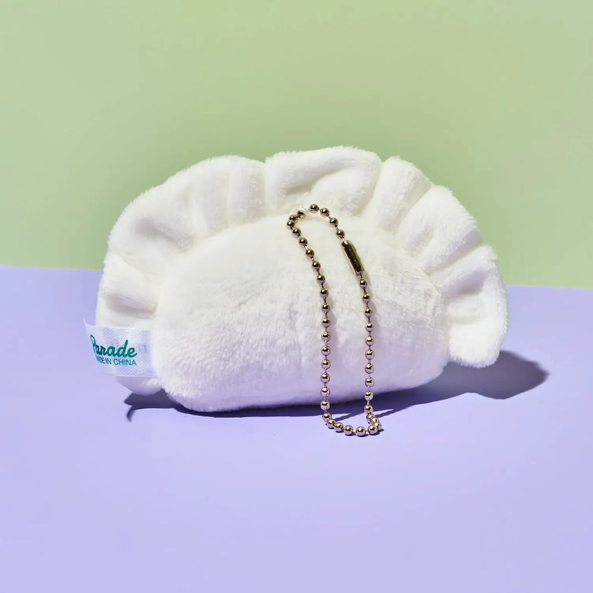 A white plush dumpling-shaped pouch with a gold ball chain strap, resting on a split pastel background of light green and lavender. A white fabric tag on the side reads 'pade with grace'.