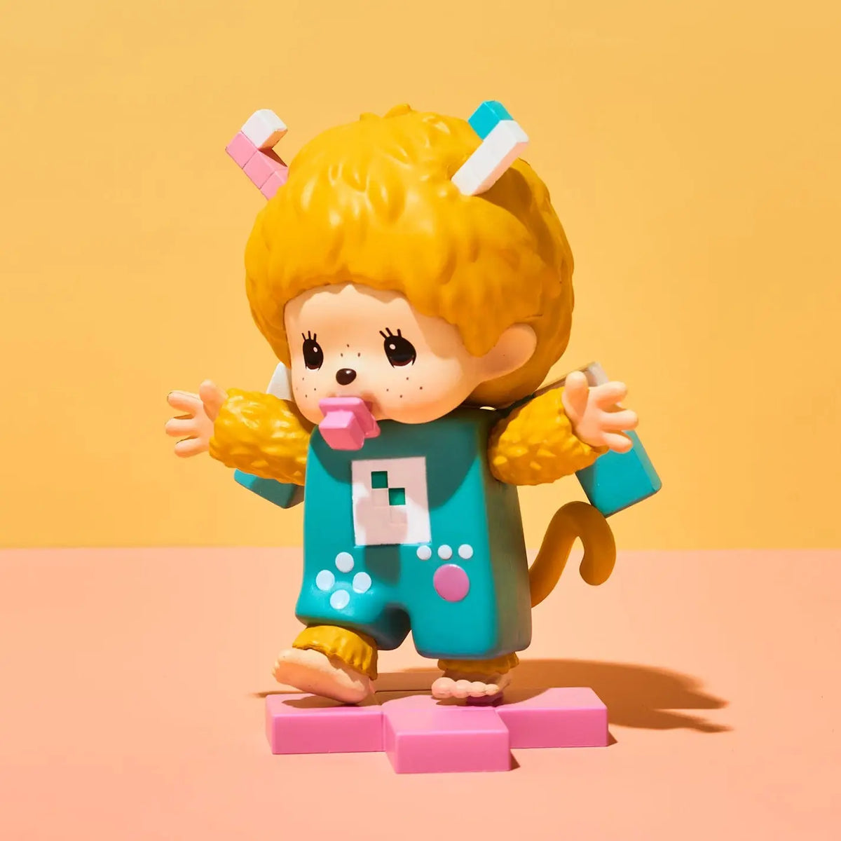 A stylized collectible figure of a child with curly yellow hair wearing a turquoise and yellow animal-inspired outfit, standing on pink blocks against a warm yellow and peach studio background.