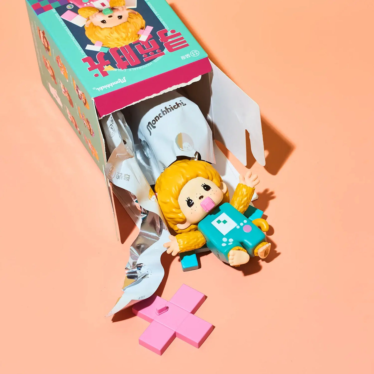 A small cartoon-style blind box figure with yellow hair and a teal outfit lies beside its open box and a pink cross-shaped accessory on a peach studio background. The packaging has colorful graphics and Chinese text, and the figure appears to be the product being sold.