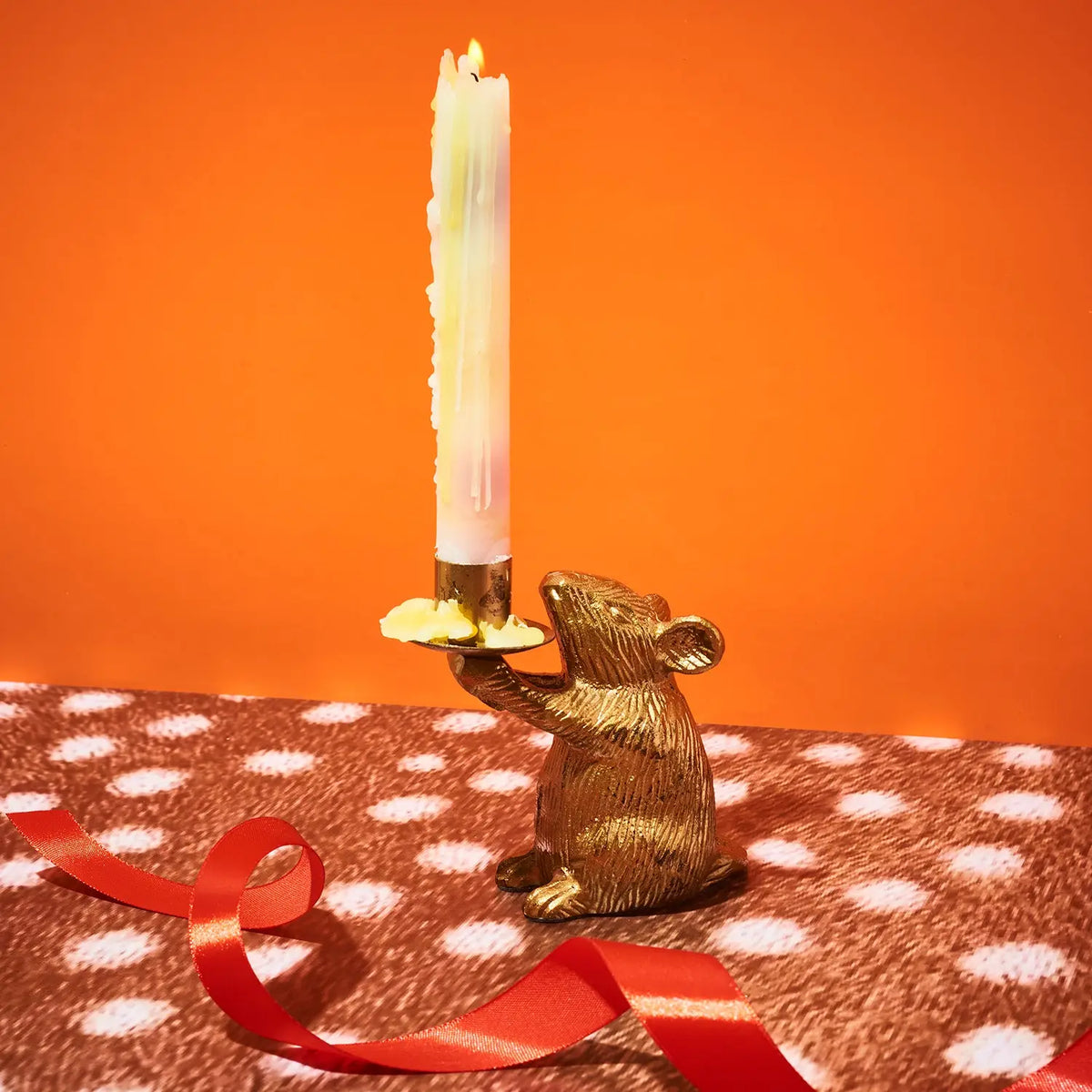A gold mouse-shaped candle holder with a lit white taper candle, set on a brown and white polka-dotted surface with a red ribbon against a solid orange background.
