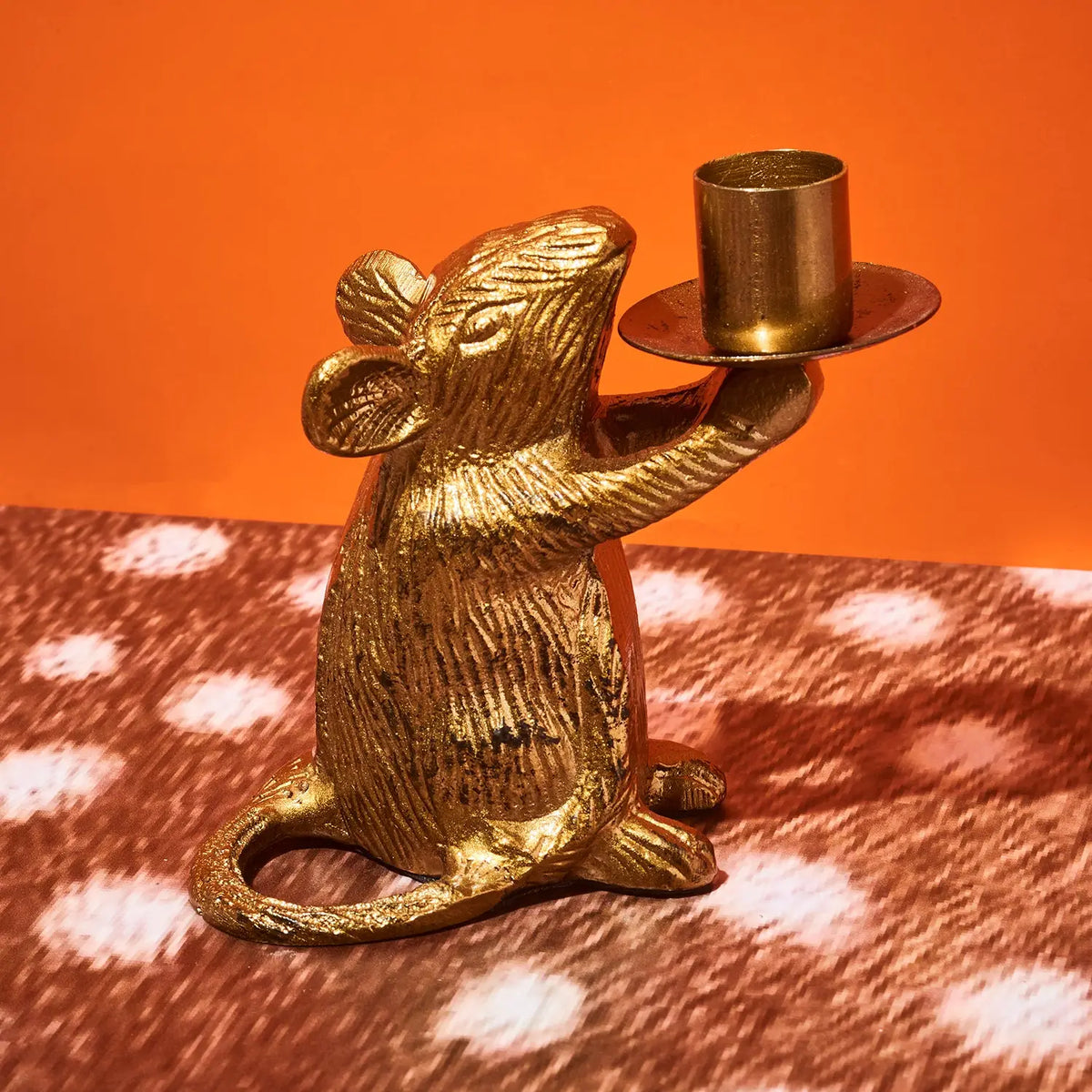 A gold-colored metal mouse figurine holding a small candle holder, set against a solid orange background and a brown-and-white polka-dotted surface.