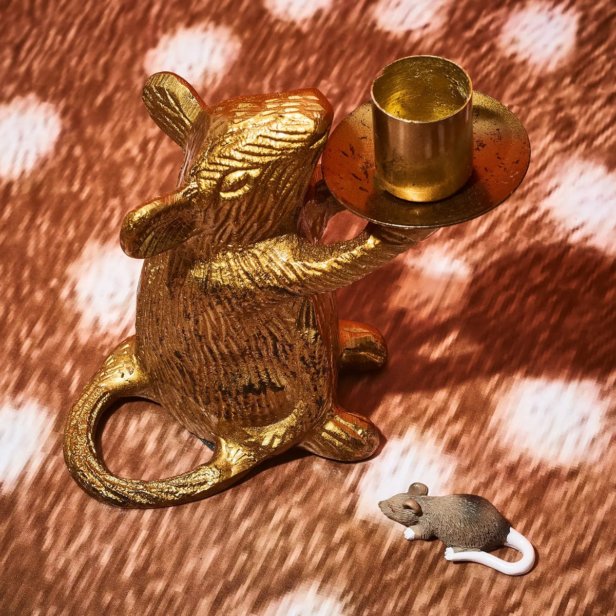 A gold-toned metal mouse figurine holding a taper candle holder, placed on a brown-and-white spotted fabric surface, with a small gray mouse figurine nearby.