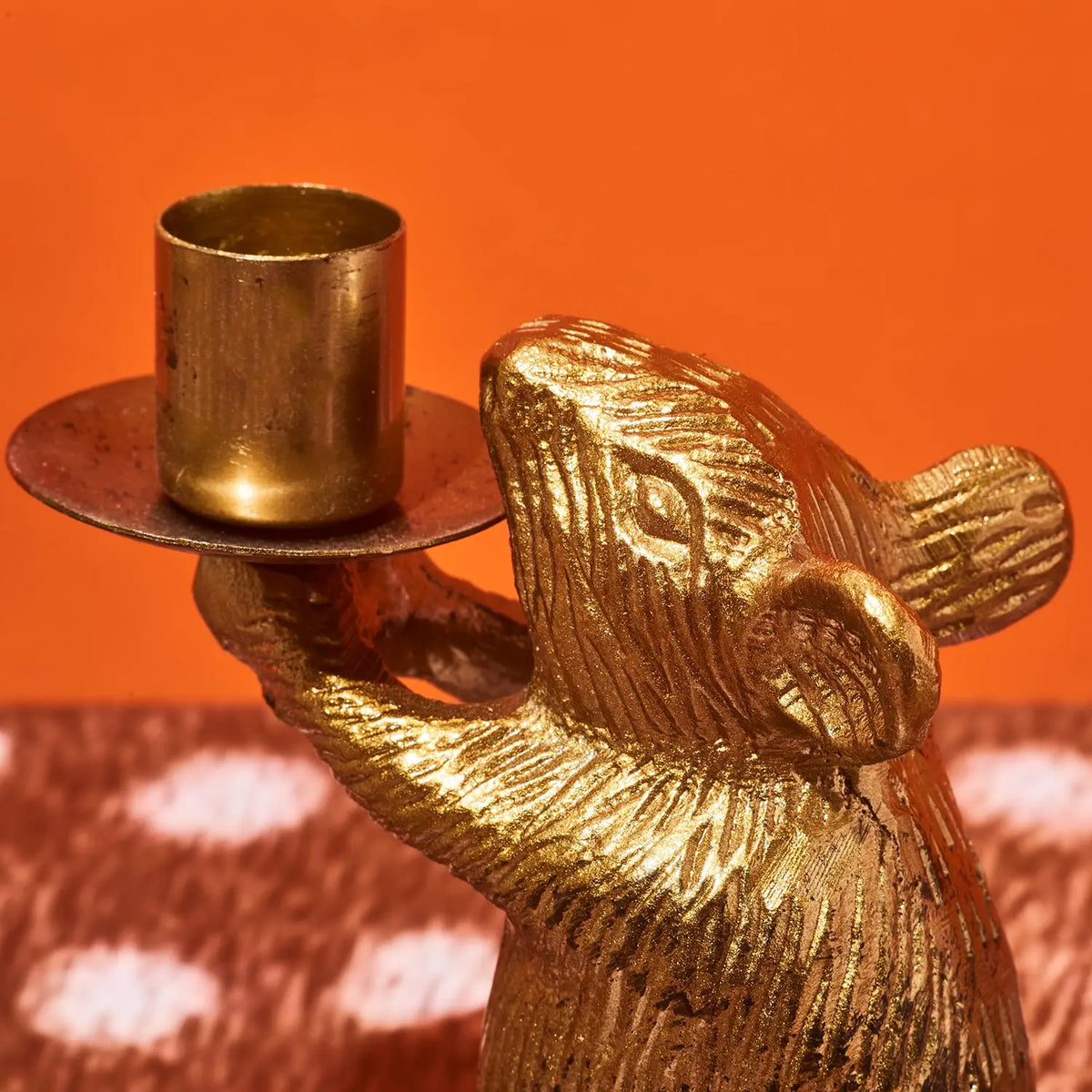A close-up of a metallic gold mouse figurine holding a small brass candle holder against a solid orange background.