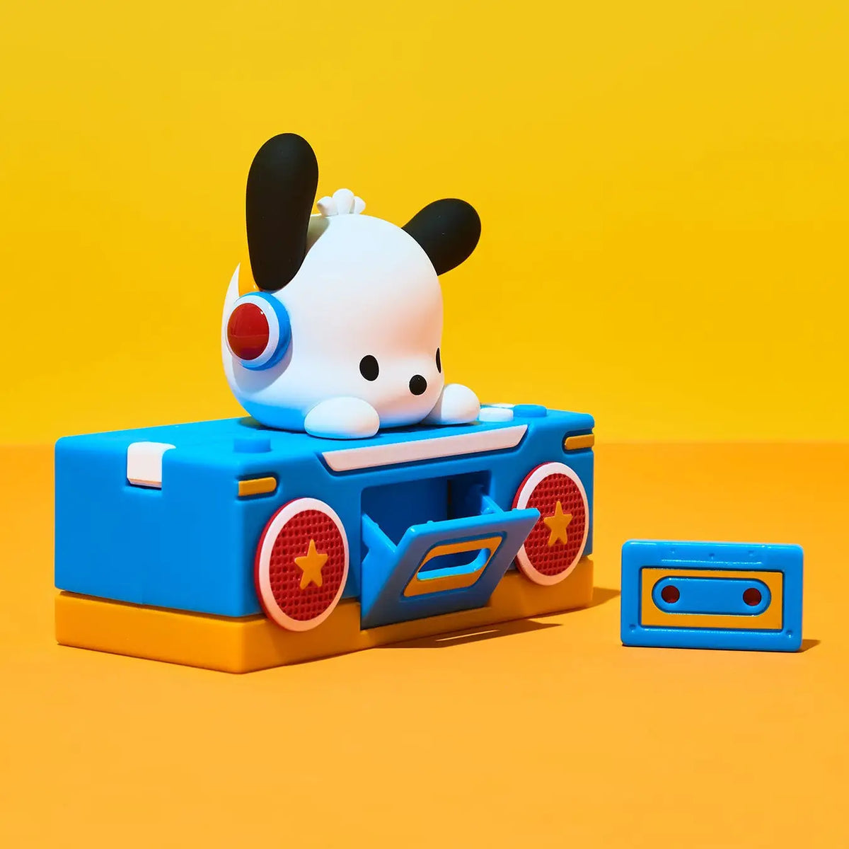 A stylized blue cassette-deck toy with a white cartoon dog character on top, shown on a bright yellow and orange studio background. A matching blue cassette sits beside it.