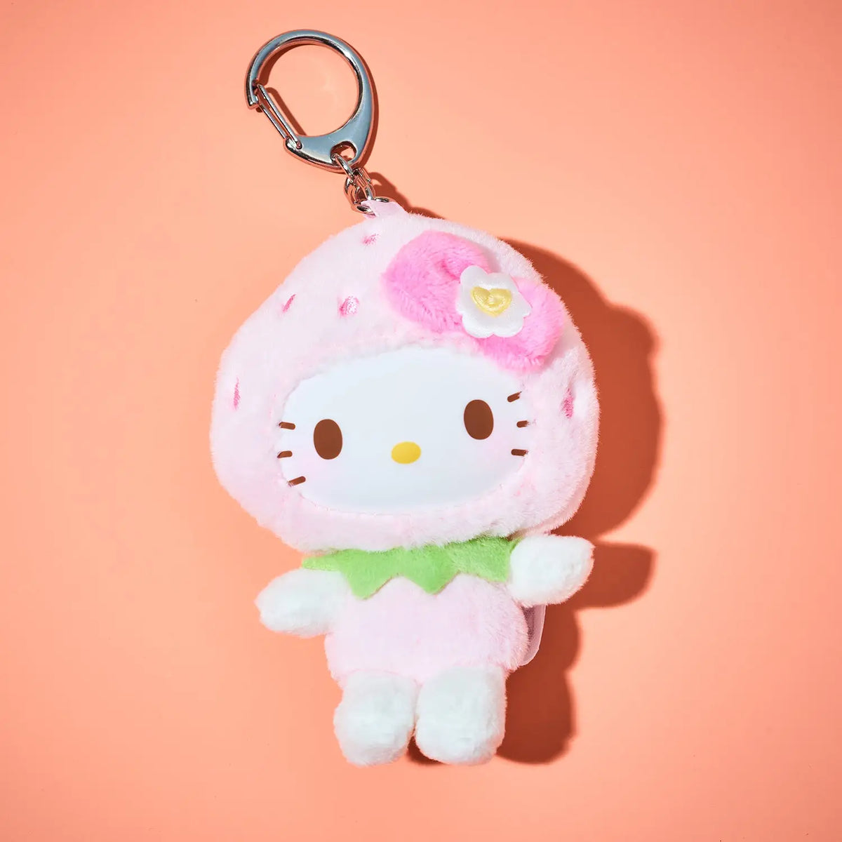 A Hello Kitty strawberry-themed plush keychain charm with a pink hood, white face, green collar, and silver clasp, set against a solid peach background.