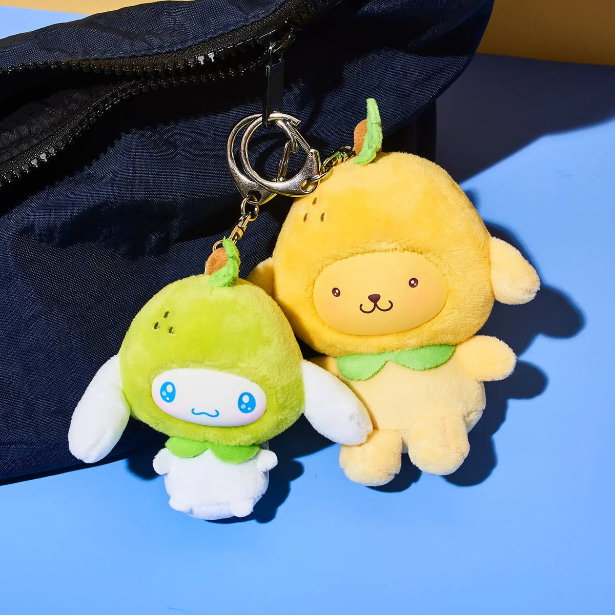 Two plush character keychains, one green and one yellow, attached to a silver metal ring, resting against a dark navy bag on a blue surface.