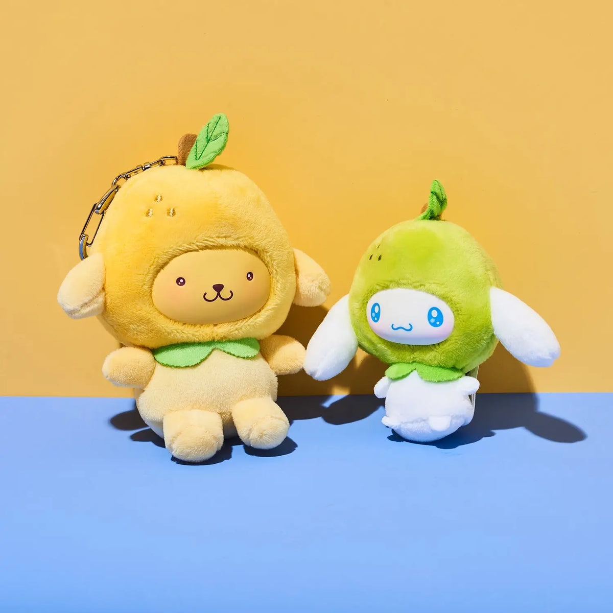 Two cute plush character keychains shaped like fruit characters, one yellow and one green, displayed on a blue surface against a yellow background.