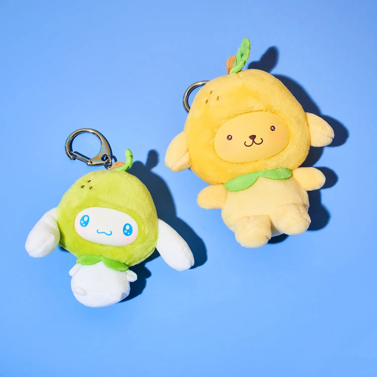 Two plush character keychains on a solid blue background. One is lime green with a white face and white wings, the other is pale yellow with a green collar and a small leaf on its head.