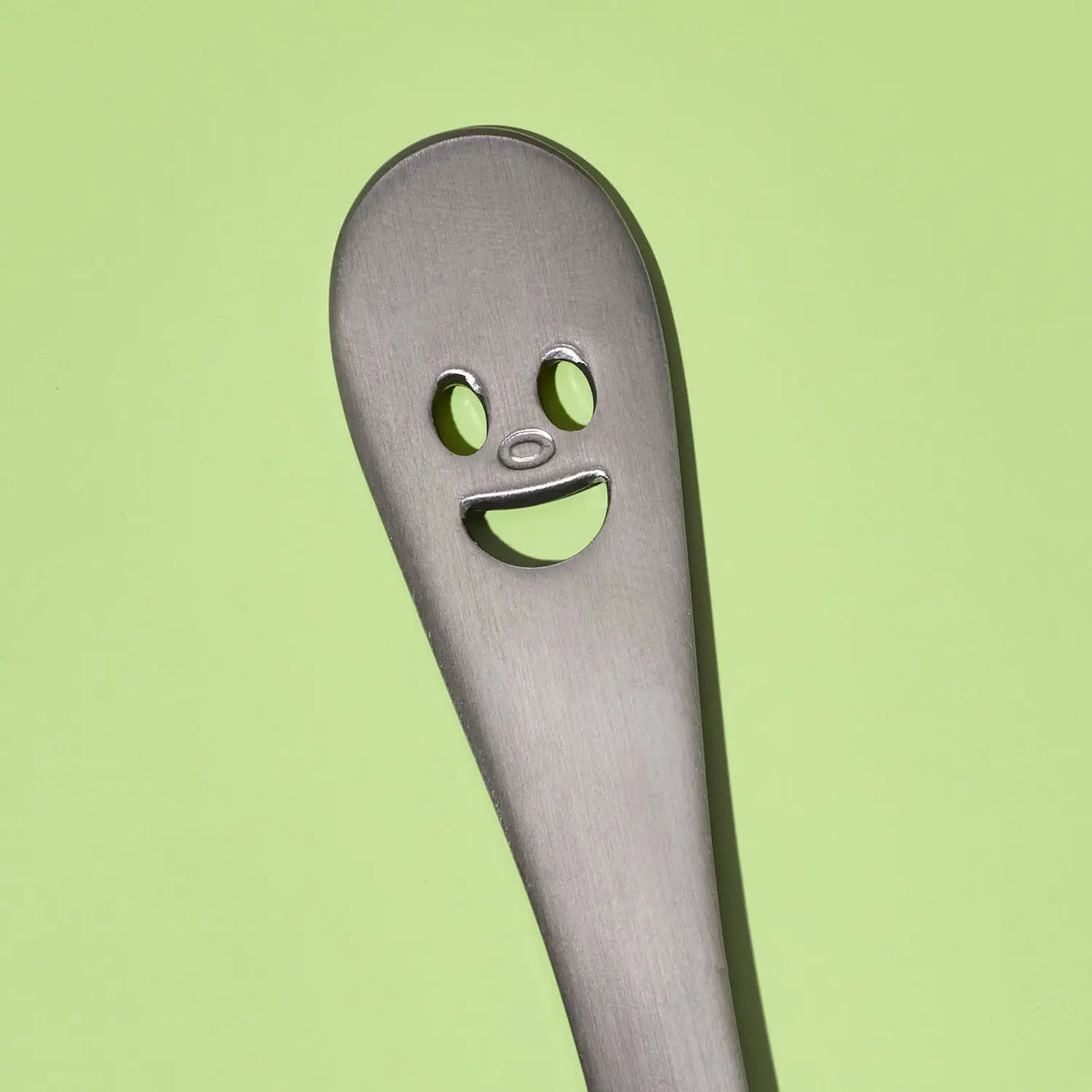 A close-up of a stainless steel utensil handle with a novelty face cutout, set against a solid light green background.